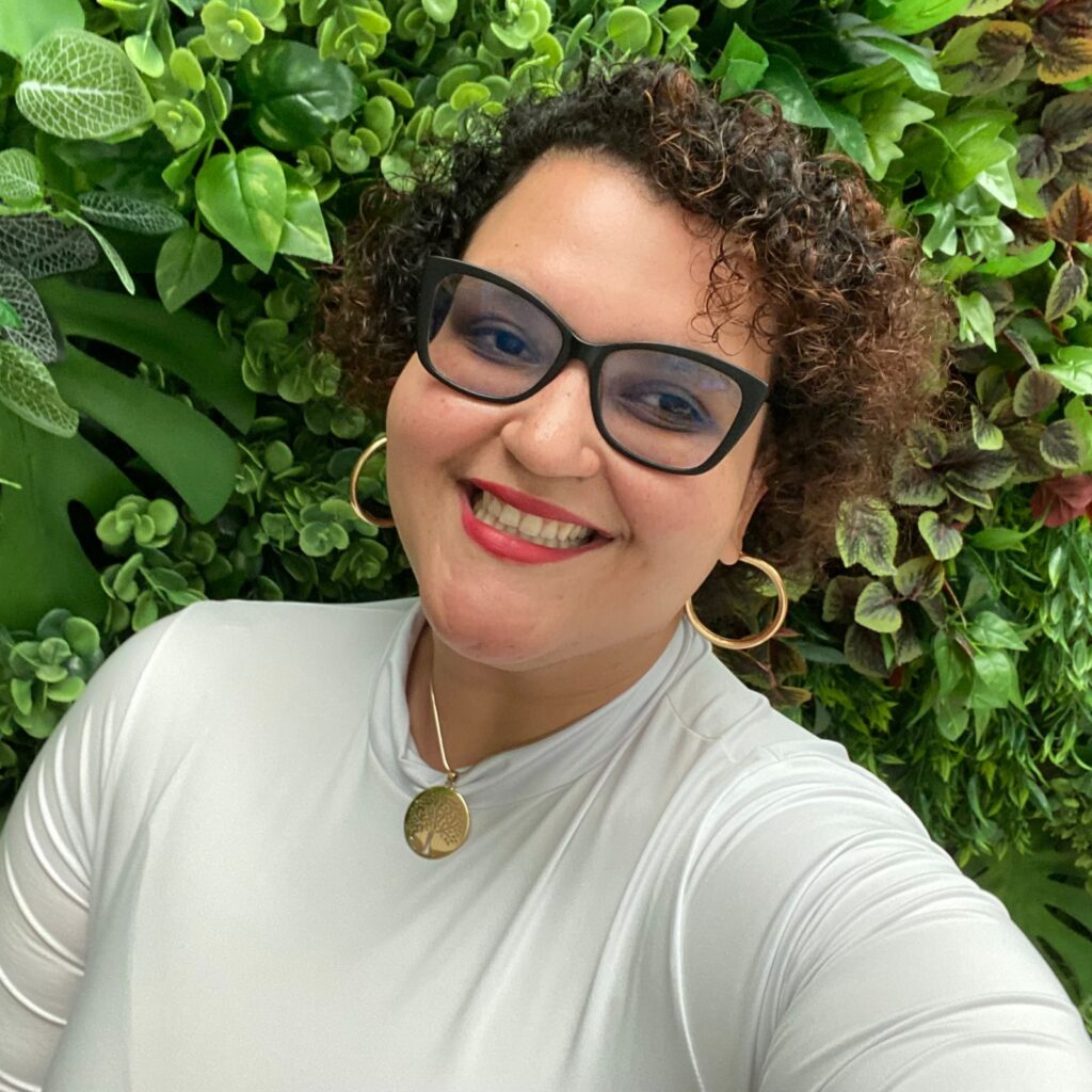 Fotografia de Alana Thomaz: mulher de pele branca, cabelos cacheados, curtos de cor castanho; veste uma blusa branca e usa óculos de grau.